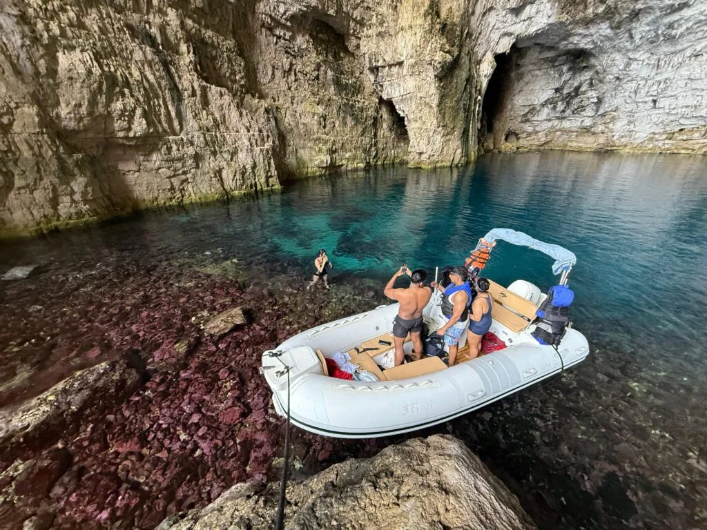 A group of people inside Haxhi Ali Cave duaring a boat trip. A small speedboat and crystal clear waters is seen.