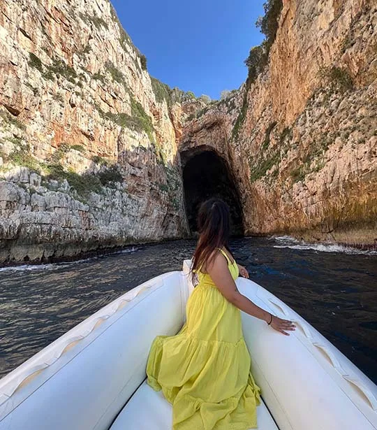 A girl, wearing a yellow dress, staying on a speed boat, posing for a photo in front of the entrance of Haxhi Ali Cave.