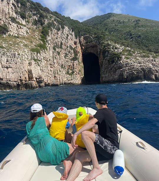 A family during a boat trip to the Haxhi Ali Cave. It is shown the entrance of the cave, Karaburun Peninsula hills and the wavy sea.